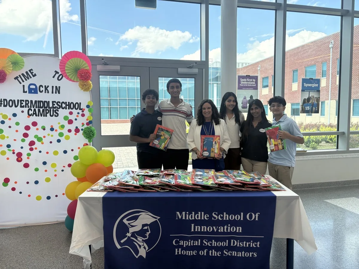 Delaware Secretary of Education Cindy Marten holding a supply kit, standing with SkillSphere's student founders behind a table of packed school supplies at the Dover Middle School campus.